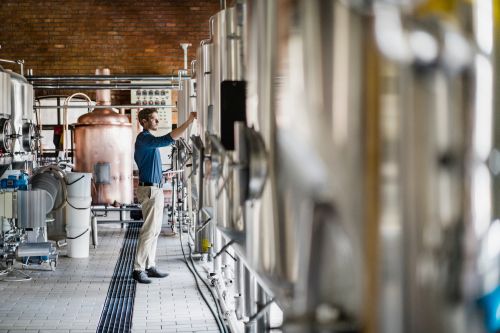 Male worker operating machines in the brewery. He holds a tablet in his hand while standing in the middle of metal kettles.